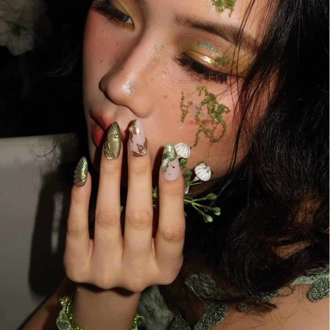 Close-up of a person with floral nail art and face paint, wearing a green dress.